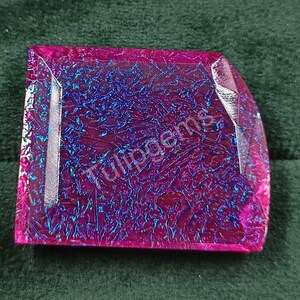 Puede incluir: Una gema iridiscente de corte cuadrado con un vibrante tono magenta y una superficie texturizada. La gema presenta vetas azules y el texto "Tulipgems". Esta gema podría usarse para joyería u otros fines decorativos.