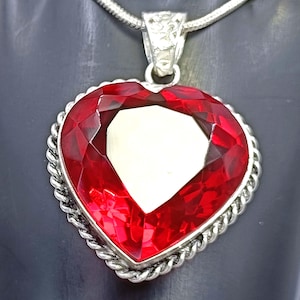 May include: A heart-shaped pendant with a large, faceted red gemstone. The gemstone is set in a silver-toned metal frame with a rope-like border. The pendant hangs from a silver chain.