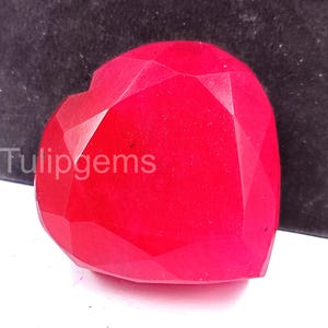 Puede incluir: Una gema facetada en forma de corazón, de color rojo vibrante. La gema tiene múltiples facetas que capturan la luz, creando un efecto brillante. El texto "Tulipgems" es visible en el lado izquierdo de la gema.