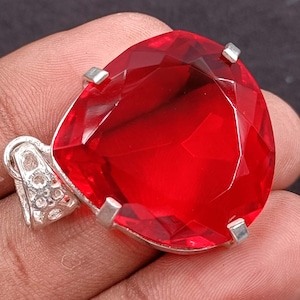 Könnte beinhalten: Ein facettierter, herzförmiger, roter Edelsteinanhänger mit einer silberfarbenen Öse. Der Edelstein ist ein leuchtendes, transparentes Rot. Der Anhänger ist mit vier silbernen Zinken gefasst. Die Öse hat ein dekoratives Design.