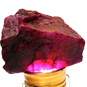 May include: A large, rough-cut ruby gemstone with a deep red hue. The raw mineral displays a textured surface and varying shades of red, with a bright pink glow emanating from within. The ruby is displayed on a gold-colored stand.