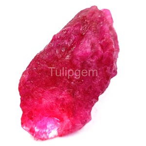 Puede incluir: Una gema de rubí en bruto, sin cortar, con un vibrante tono rosa-rojizo. La piedra natural tiene una forma irregular y una superficie texturizada. El texto "Tulipgem" es visible en la superficie de la gema.