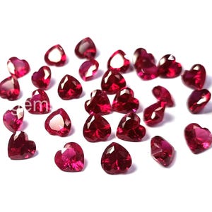 May include: A collection of heart-shaped, faceted gemstones in a deep red hue. The gemstones are scattered across a white surface, with varying sizes and angles, creating a sparkling effect. Ideal for jewelry making.