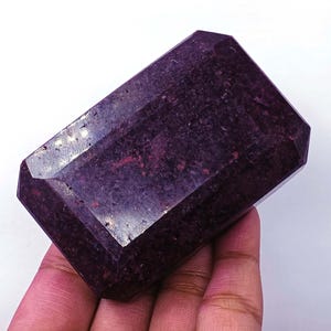 Puede incluir: Una gema rectangular grande de color burdeos intenso. La gema tiene múltiples facetas y una superficie pulida que refleja la luz. La piedra se sostiene en una mano, mostrando su tamaño y forma. La gema parece ser un rubí o una piedra preciosa similar.