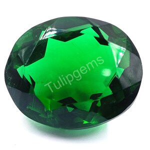 May include: A faceted, oval-shaped, emerald-green gemstone. The gem has a high degree of clarity and brilliance, with multiple facets reflecting light. The text "Tulipgems" is printed on the surface of the gem.