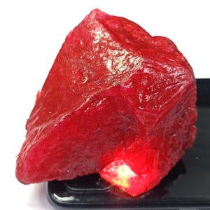 May include: A large, rough-cut ruby gemstone with a deep red color. The raw mineral has a textured surface and is resting on a black surface. The ruby is a natural, uncut gemstone.