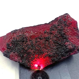 Puede incluir: Una gema de rubí en bruto y sin cortar, de color rojo intenso y superficie texturizada. El rubí presenta parches de depósitos minerales negros, y una luz roja brillante emana de la parte inferior. Esta piedra preciosa natural es ideal para la fabricación de joyas o coleccionar.