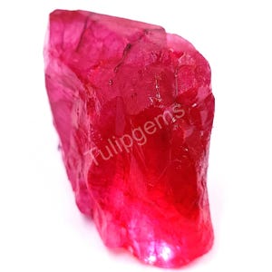 Peut inclure: Une pierre précieuse de rubis brute et non taillée, d'un rouge vif. La surface rugueuse de la pierre est visible, avec le texte "Tulipgems" imprimé sur le côté. Le rubis est posé sur un fond blanc.