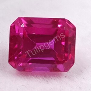 Può includere: Una gemma magenta sfaccettata a taglio rettangolare. La gemma ha una superficie lucida ed è posta su uno sfondo bianco. Il testo "Tulipgems" è visibile sulla superficie della gemma.