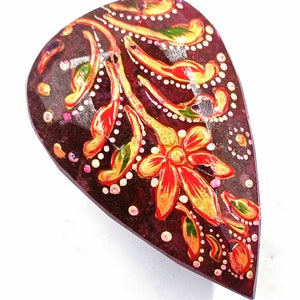 May include: A teardrop-shaped, hand-painted wooden object with a dark burgundy base. The surface is adorned with a floral design in red, gold, and green, accented with white dots. The object has a glossy finish.