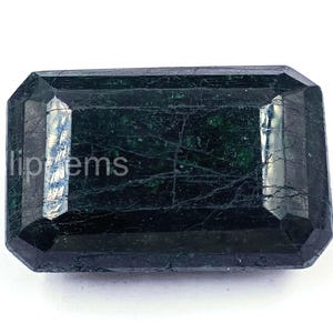 Puede incluir: Una piedra preciosa de esmeralda rectangular de color verde oscuro con un corte facetado. La gema tiene un color profundo y rico e inclusiones internas visibles. El texto "lipgems" es visible.
