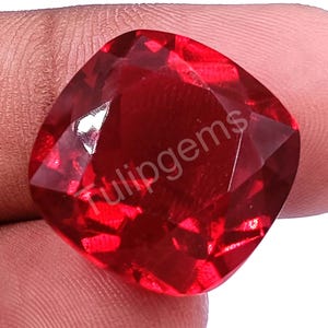 Op de afbeelding: Een gefacetteerde, kussenvormige edelsteen in een dieprode tint. De edelsteen heeft een hoge mate van helderheid en glans, met meerdere facetten die het licht vangen. De tekst "Tulipgems" is zichtbaar.