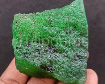 May include: A rough-cut, vibrant green emerald gemstone. The surface of the emerald is textured and irregular, with areas of darker green and black. The text "tulipgems" is visible on the surface. The gemstone is held in a hand.