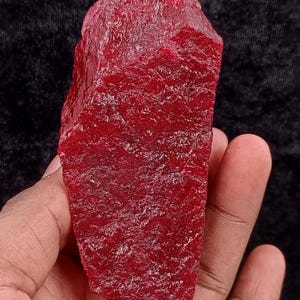 May include: A rough-cut, deep red gemstone with a textured surface. The ruby is held in a hand against a dark background. The text "Tulipgems" is visible at the bottom of the image.