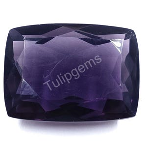 May include: A faceted, rectangular-shaped gemstone with a deep purple hue. The gem has a polished surface with visible internal inclusions and the text "Tulipgems" printed on the surface.