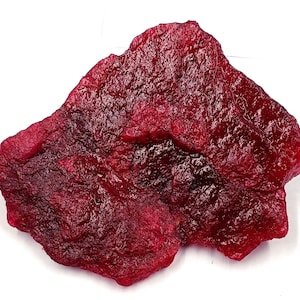 May include: A rough-cut, irregularly shaped gemstone in a deep, rich red hue. The surface has a textured appearance, with variations in color and tone. The stone is displayed against a white background, highlighting its natural beauty.