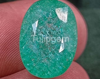 Pode incluir: Gema de esmeralda oval e facetada. A gema é de cor verde vibrante com inclusões internas e facetas refletoras. O texto "Tulipgem" é visível na superfície da pedra.