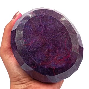 May include: A large, faceted gemstone with a deep purple hue, held in a hand. The gem's surface displays internal patterns and a polished, reflective quality. The cut of the stone is multi-faceted.