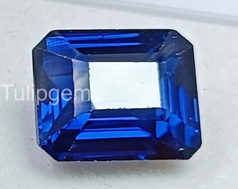 May include: A faceted, emerald-cut sapphire gemstone. The gem is a deep, vibrant blue with a rectangular shape and beveled edges. The word "Tulipgem" is visible in the upper left corner. The gem's facets reflect light, creating a sparkling effect.