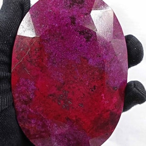 May include: A large, oval-shaped ruby gemstone with a faceted surface. The gem displays a deep red and magenta color with variations in tone. The stone is held by a gloved hand against a white background, highlighting its polished facets and natural inclusions.