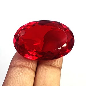 以下が含まれることがあります： 鮮やかで半透明な赤色をした、大きな楕円形のファセットカット宝石。石の表面には複雑な幾何学模様のカットが施されており、光を捉えて反射する多数のファセットが全体に広がっています。