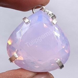 May include: A pear-shaped, faceted gemstone pendant with a soft, opalescent pink hue. The pendant is held in place by silver-colored metal prongs and a loop at the top. The text "Tulipgems" is visible on the surface.