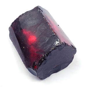 May include: A raw, uncut gemstone with a deep burgundy color and a rough, textured surface. The stone has a rectangular shape with visible facets and a polished, reflective interior. The gemstone is set against a white background.