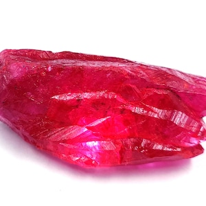 Puede incluir: Una gema de rubí en bruto, sin cortar, de color rojo intenso. La superficie de la piedra tiene un aspecto rugoso y texturizado con facetas visibles. La gema se muestra sobre un fondo blanco, resaltando su color y forma.