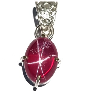 May include: A silver pendant with a large, oval, deep red gemstone. The gemstone has a star-like pattern across its surface. The pendant's bail is intricately designed, and the setting has four prongs holding the stone.