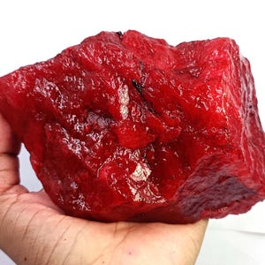 May include: A large, raw, irregularly shaped mineral specimen with a vibrant, deep red colour. The surface of the stone features a glossy, crystalline texture with visible facets, natural ridges, and small dark inclusions throughout the structure.
