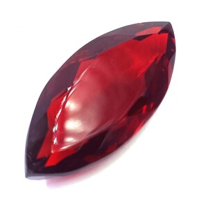 Può includere: Una pietra preziosa con taglio a marquise di colore rosso profondo e traslucido. La pietra presenta molteplici sfaccettature triangolari e trapezoidali sulla superficie, creando un aspetto lucido e riflettente con estremità appuntite.