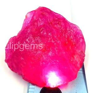 Puede incluir: Una gema grande, en bruto, de color rosa intenso, con una superficie texturizada. La gema tiene una forma natural e irregular y un color vibrante y saturado. La palabra "ulipgems" es visible en la superficie de la gema.