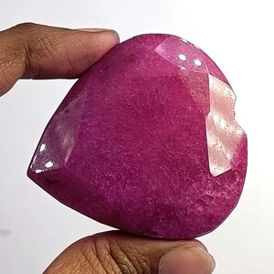 Pode incluir: Uma grande pedra preciosa facetada em forma de coração, em um tom magenta profundo. A pedra tem uma superfície polida com texturas internas visíveis e algumas imperfeições. A gema é segurada entre dois dedos contra um fundo branco.