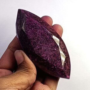 Puede incluir: Una gema facetada y alargada de color púrpura intenso. La gema tiene múltiples facetas pulidas que reflejan la luz, creando un efecto brillante. La forma de la piedra es similar a un corte marquesa, con extremos puntiagudos y un centro ancho.