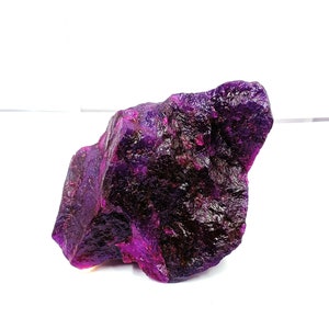 May include: A raw, uncut gemstone with a deep purple hue. The surface of the stone has a rough, textured appearance, with areas of lighter and darker shades of purple. The gemstone is set against a white background, highlighting its natural beauty.