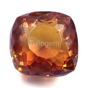 Puede incluir: Una gema facetada, de talla cojín, con tonos naranja y marrón. La gema tiene un núcleo dorado que se funde en bordes marrón rojizo. El texto "Tulipgem" es visible en la superficie. La gema está sobre un fondo blanco.