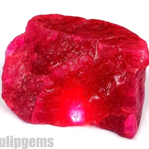 Può includere: Una gemma di rubino grezza, non tagliata, di un colore rosso vibrante. La pietra naturale ha una superficie strutturata e una forma irregolare, che mostra il suo stato grezzo e non lucidato. L'immagine è di una gemma, probabilmente per gioielli o collezioni.