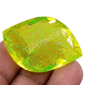 Puede incluir: Una gema grande y facetada con un vibrante tono amarillo-verdoso. La gema tiene una forma irregular única y una superficie pulida que refleja la luz. El texto "Tulipgems" es visible en la superficie de la gema.