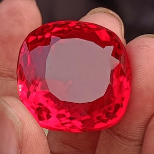 May include: A large, faceted, cushion-cut gemstone in a vibrant red colour. The gem has a high degree of clarity, reflecting light across its many facets. The gemstone is held between fingers, showcasing its size and brilliance. This could be a ruby or a similar precious stone.