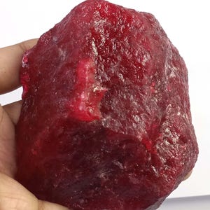 Könnte beinhalten: Ein roher, ungeschliffener Rubin-Edelstein mit tiefroter Farbe. Die Oberfläche des Steins hat ein strukturiertes Aussehen mit Farbvariationen und Lichtreflexionen. Der Rubin wird in einer Hand gehalten, um seine Größe und natürliche Form zu zeigen.