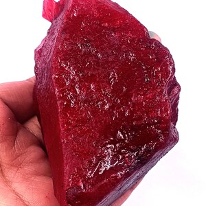 Könnte beinhalten: Ein großer, ungeschliffener Rubin-Edelstein mit tiefroter Farbe. Das rohe Mineral hat eine strukturierte Oberfläche und eine unregelmäßige Form, die in einer Hand vor weißem Hintergrund gehalten wird. Der Rubin scheint ungeschnitten und natürlich zu sein.