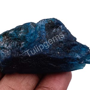Puede incluir: Una gema en bruto, sin cortar, de color verde azulado intenso. La piedra tiene una superficie texturizada y una forma irregular. El texto "Tulipgems" es visible en la superficie. La gema se sostiene en una mano.