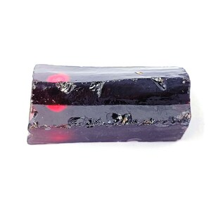 Puede incluir: Una gema oscura y rectangular con una apariencia en capas. La capa superior es de un negro profundo y opaco, mientras que el interior revela vetas de rojo vibrante. Los bordes tienen una textura rugosa y natural, sobre un fondo blanco.