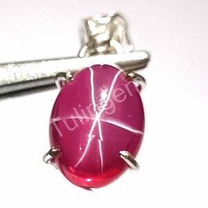 May include: A polished, oval ruby gemstone with a star pattern. The deep red gem has white lines radiating from the centre. It is held by a silver metal setting. The text "Tulingen" is visible on the gem.