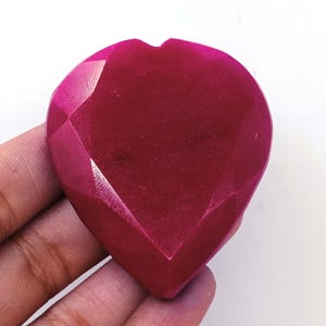 Puede incluir: Una gema facetada en forma de corazón, de un tono magenta intenso. La piedra tiene una superficie pulida con múltiples facetas, creando un juego de luces y sombras. La gema se sostiene en una mano sobre un fondo blanco.
