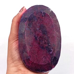 May include: A large, oval-shaped gemstone with a deep red and purple color. The gem is held in a hand, showcasing its size and faceted surface. The stone has a polished appearance, reflecting light and displaying internal patterns.