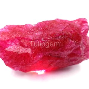 Puede incluir: Una gema de rubí en bruto y sin cortar, de color rojo vibrante. La piedra natural tiene una superficie texturizada y una forma irregular. La imagen incluye el texto "Tulipgem".