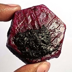 May include: A rough-cut ruby gemstone, held between fingers, displays a deep red hue with a dark, almost black, inclusion. The surface has a textured appearance with natural facets. The gemstone is irregularly shaped.