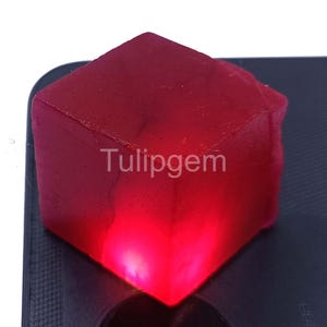 Może przedstawiać: Przezroczysty, rubinowo-czerwony sześcian z kamienia szlachetnego z napisem "Tulipgem" na powierzchni. Sześcian jest podświetlony od wewnątrz, tworząc efekt świecenia. Kamień szlachetny jest umieszczony na ciemnym tle.