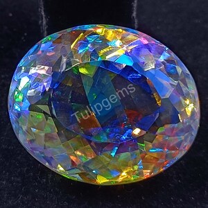 Puede incluir: Una gema grande y facetada de forma redonda. La gema muestra una vibrante gama de colores, incluyendo azul, verde, amarillo y naranja, creando un efecto arcoíris. El texto "Tulipgems" es visible. La gema está sobre un fondo oscuro.